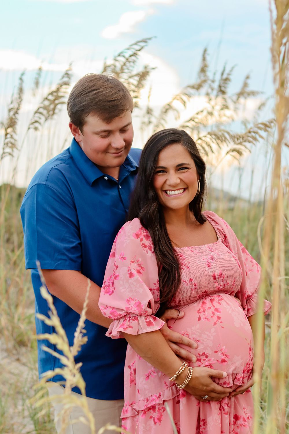 Couple poses among beach grass as they celebrate expecting a baby, wearing blue and pink florals.