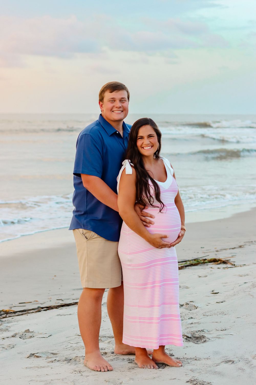 Expecting couple shares tender moment on beach at sunset wearing coordinating blue and pink outfits.