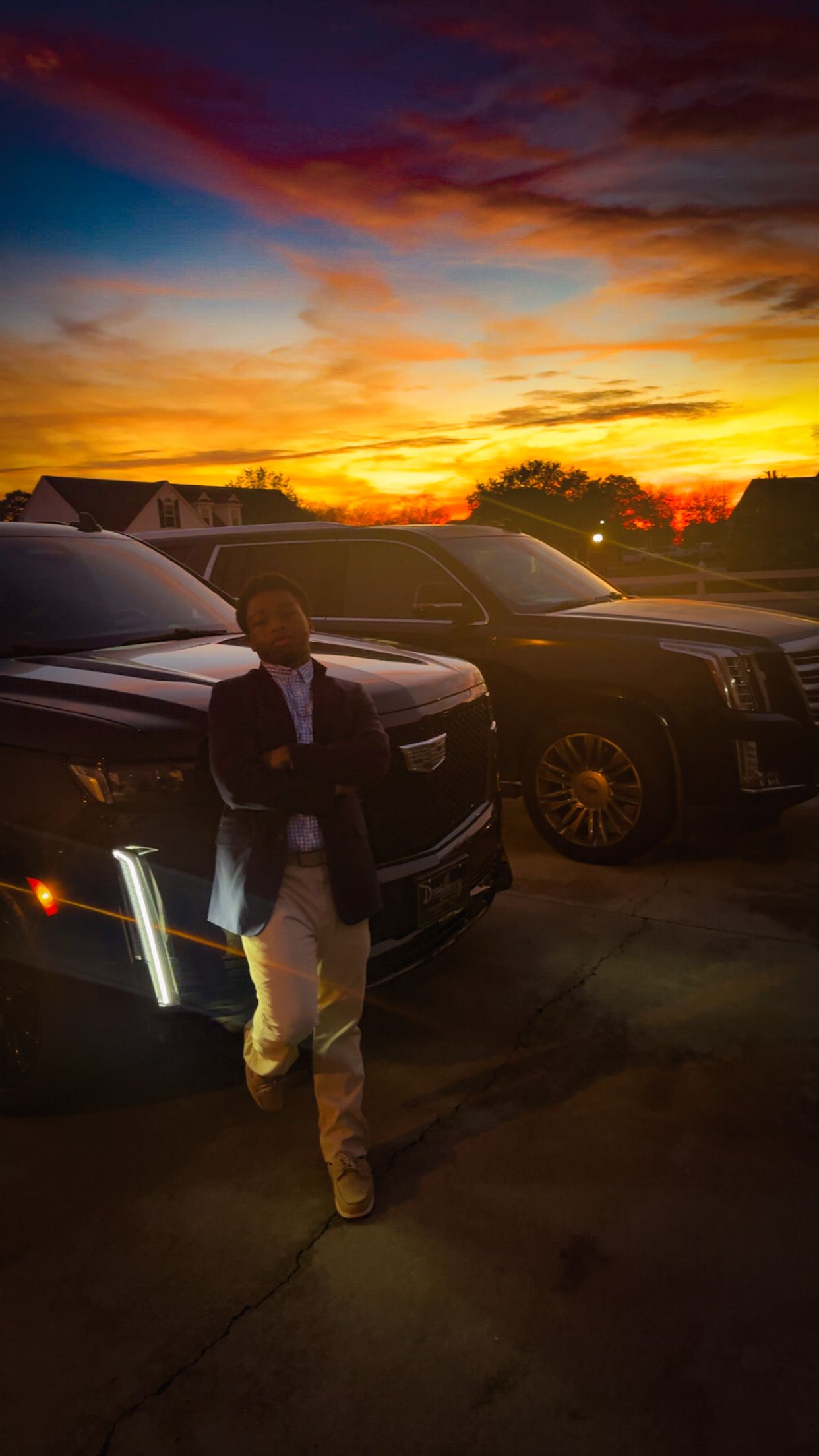 Man in suit leaning against black Cadillac Escalade SUV at golden sunset.