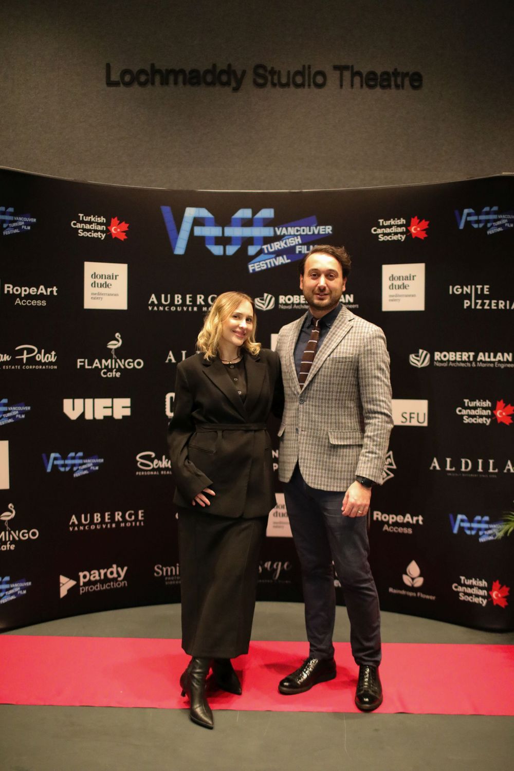 Two people pose on red carpet at Loohmaddy Studio Theatre with event sponsor logos displayed on backdrop wall.