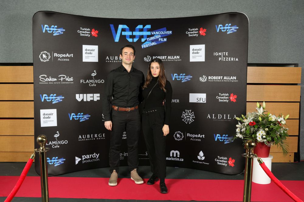 Two people in black attire pose together on a red carpet against a backdrop with sponsor logos at a formal event.