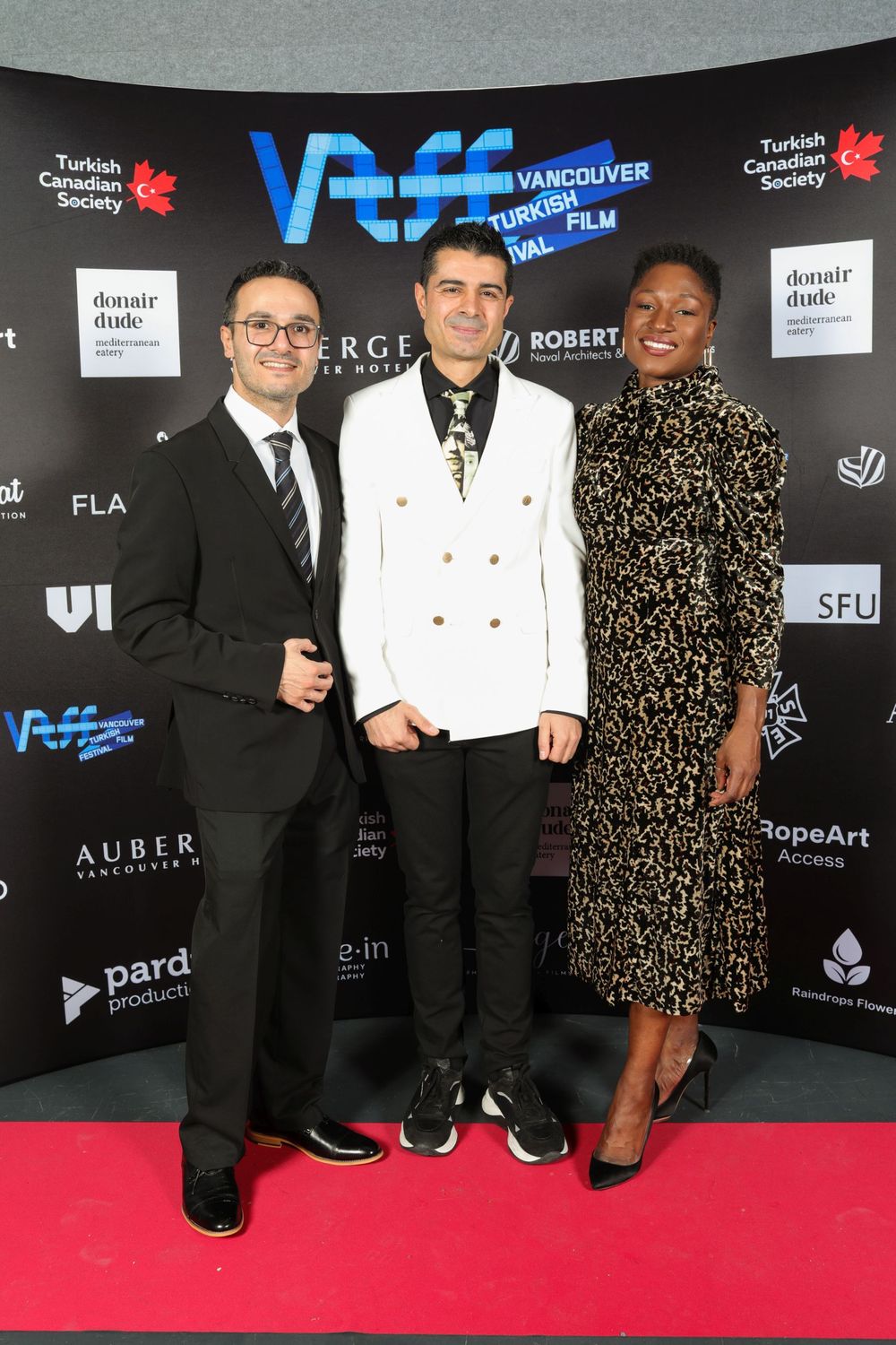 Three people pose on a red carpet at the Vancouver Turkish Film Festival with event sponsors and logos in background.