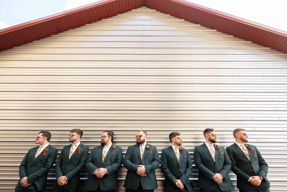 Seven groomsmen in matching dark suits lean against white siding wall during outdoor wedding photo session.