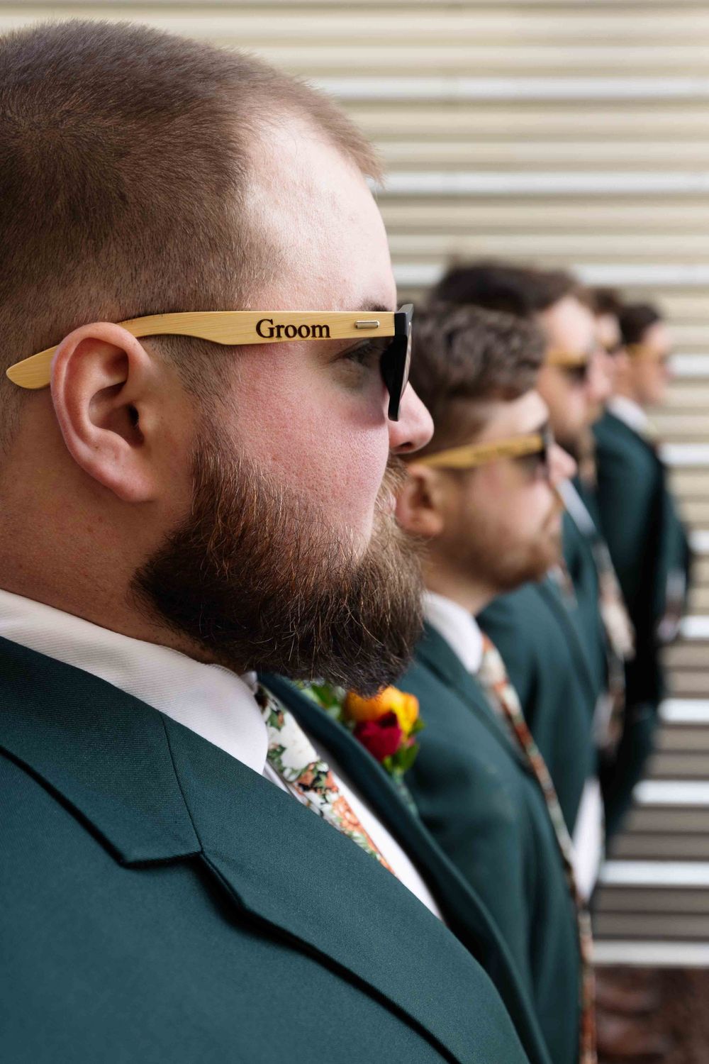 Wedding party member in a green suit wearing wooden sunglasses labeled 'Groom' stands in line during ceremony.