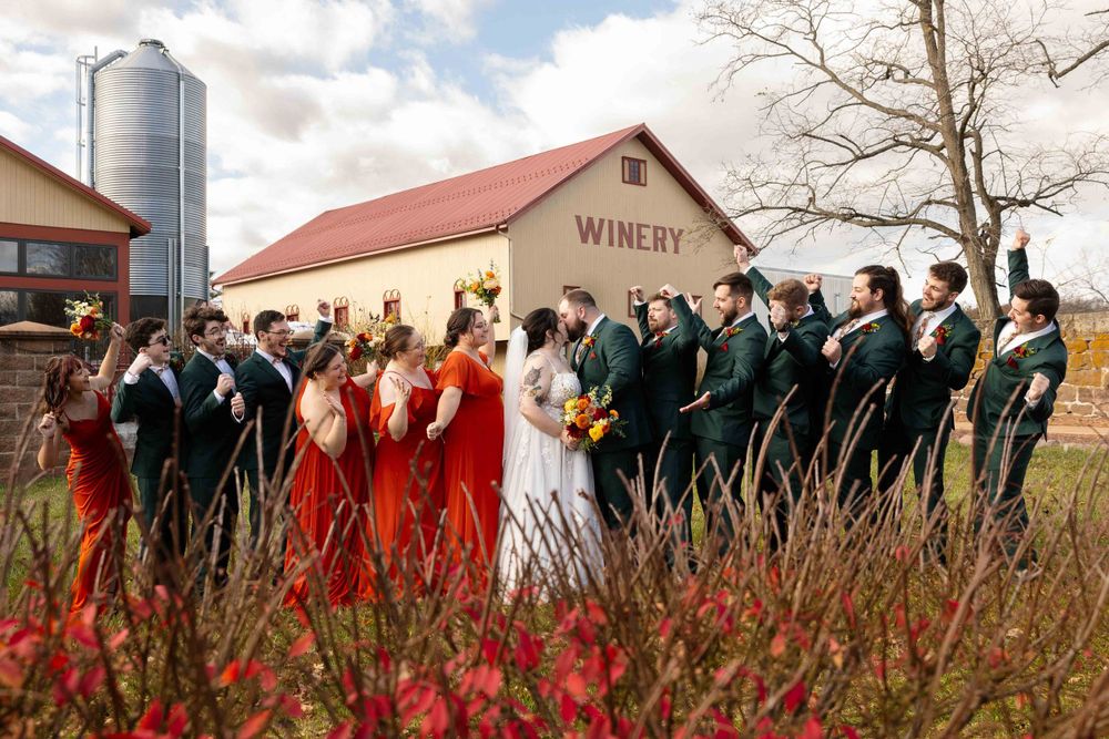 A wedding party poses together outside a rustic winery building with red flowers in the foreground.