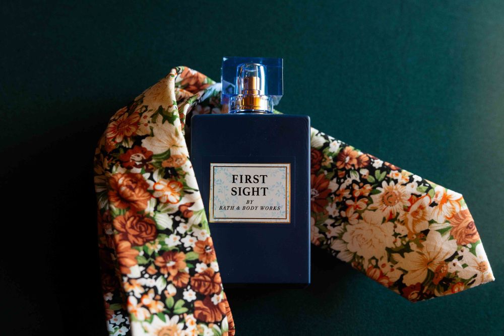 Navy blue perfume bottle labeled 'First Sight' with floral patterned fabric on dark teal background.