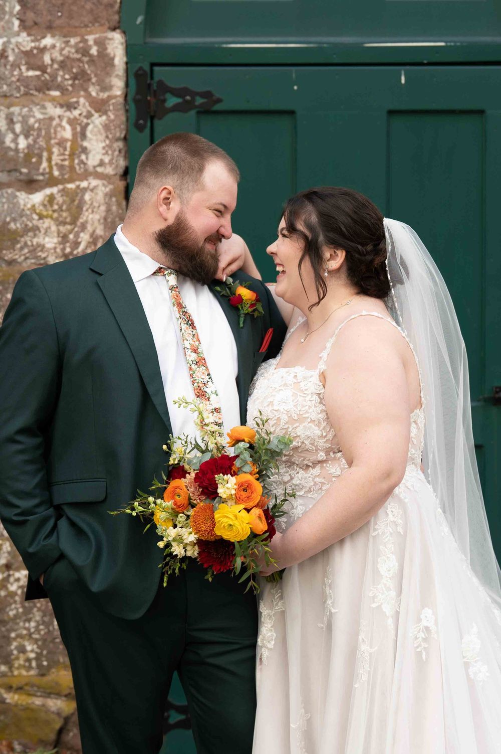 A happy wedding couple sharing a joyful moment against a green door, with the bride holding a vibrant autumn bouquet.