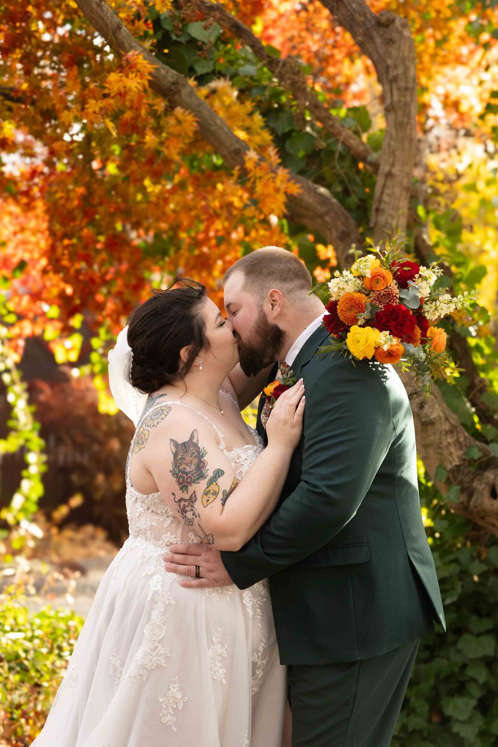 A romantic autumn wedding moment captured under vibrant orange foliage with a colorful floral bouquet in view.