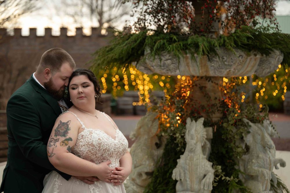 A couple shares an intimate moment under a garden fountain adorned with twinkling holiday lights at dusk.