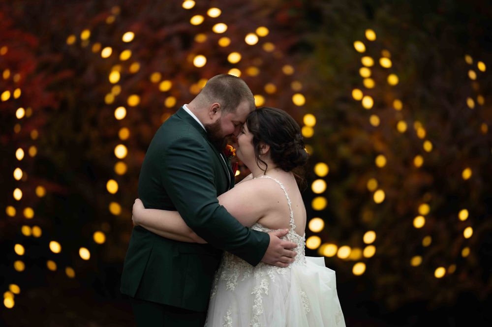 A romantic wedding couple embraces under glowing string lights in an evening garden setting.