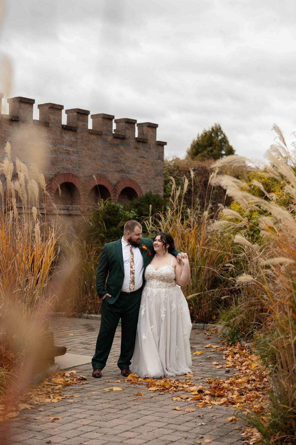 A couple in wedding attire poses in front of a castle-like stone wall surrounded by autumn foliage and ornamental grasses.