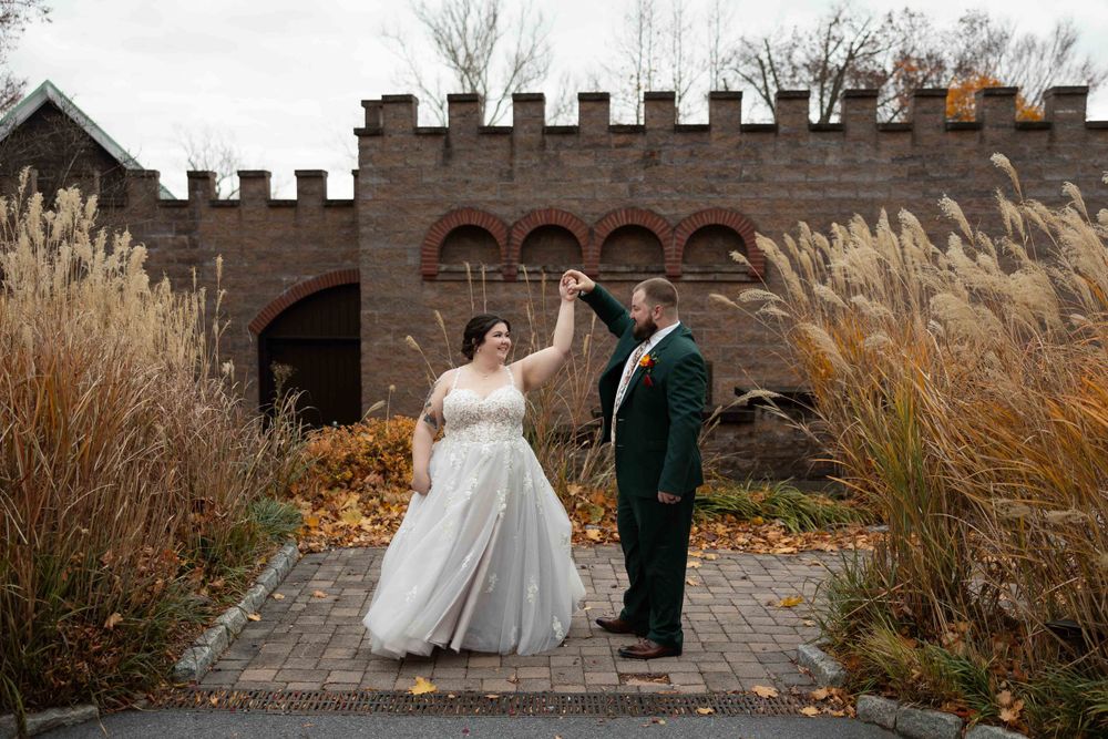 Newlyweds stand together on stone pathway in front of medieval castle wall surrounded by autumn grasses.