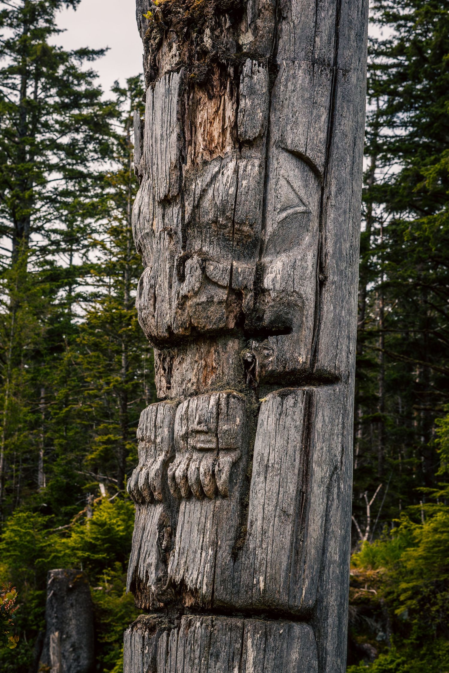 Wooden Nuxalk carving of figure wearing traditional woven hat stands near riverbank in Bella Coola, British Columbia.
