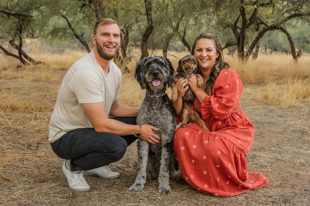 A couple in a coral dress and white shirt pose outdoors with their large gray dog during a photo session.
