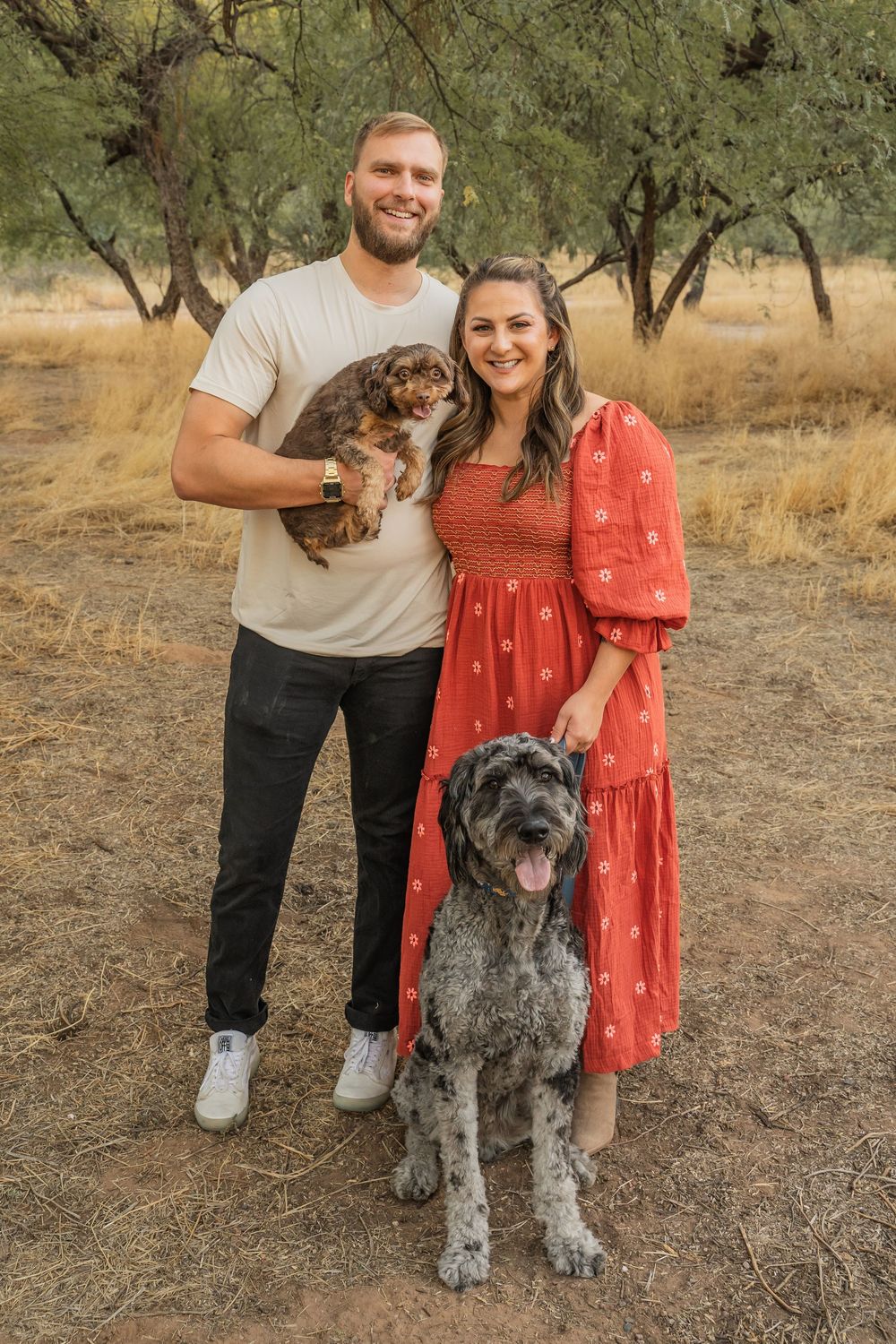 A couple poses outdoors with their two dogs - a small brown one and a large gray one - among olive trees in a rustic setting.