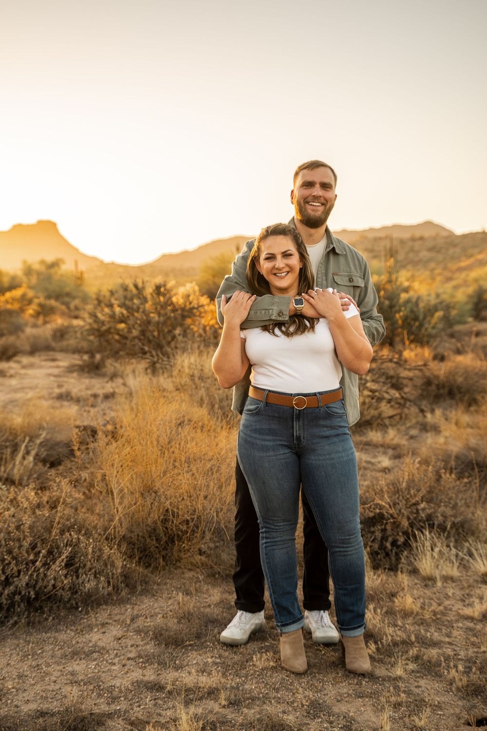 A couple poses together in a desert landscape at sunset with mountains and desert brush in the background.