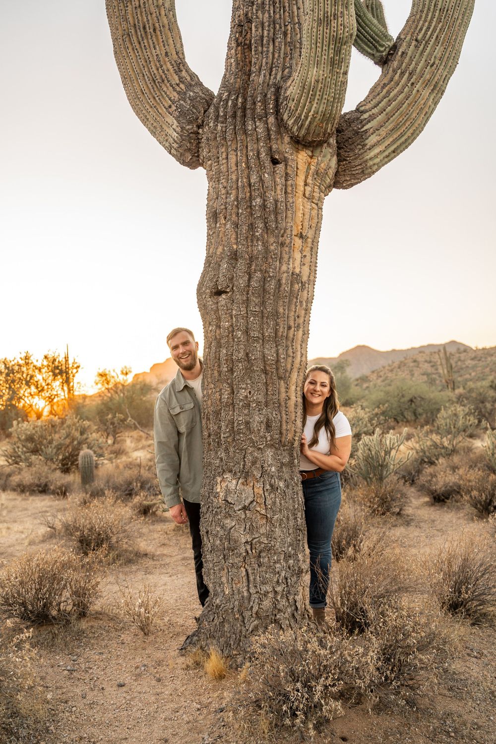Playful desert engagement photos with couple peeking around giant saguaro cactus at sunset in Arizona.