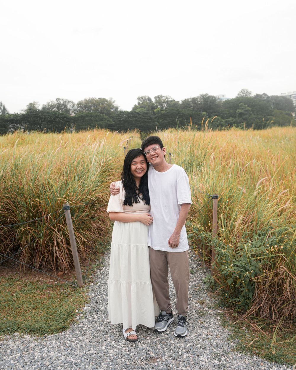 A couple stands together on a gravel path with golden grass in the background.