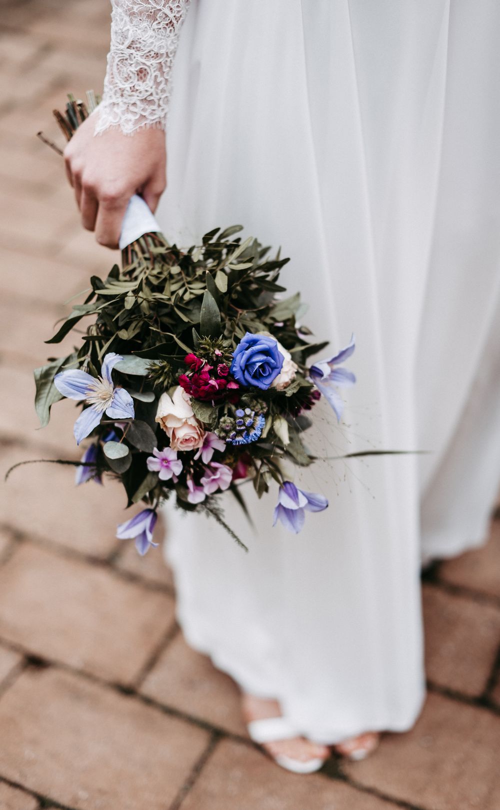 Braut in weißem Kleid hält einen Blumenstrauß mit blauen, lila und roten Blumen auf einem gepflasterten Weg.