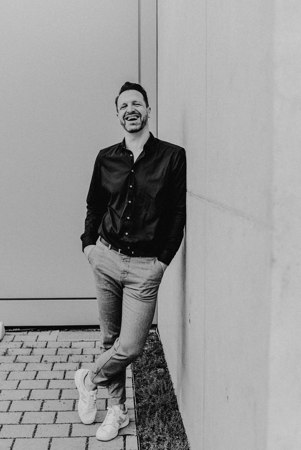 Ein lachender Mann steht angelehnt an einer Wand, der Boden ist aus grobem Stein. Schwarz-Weiss Foto.