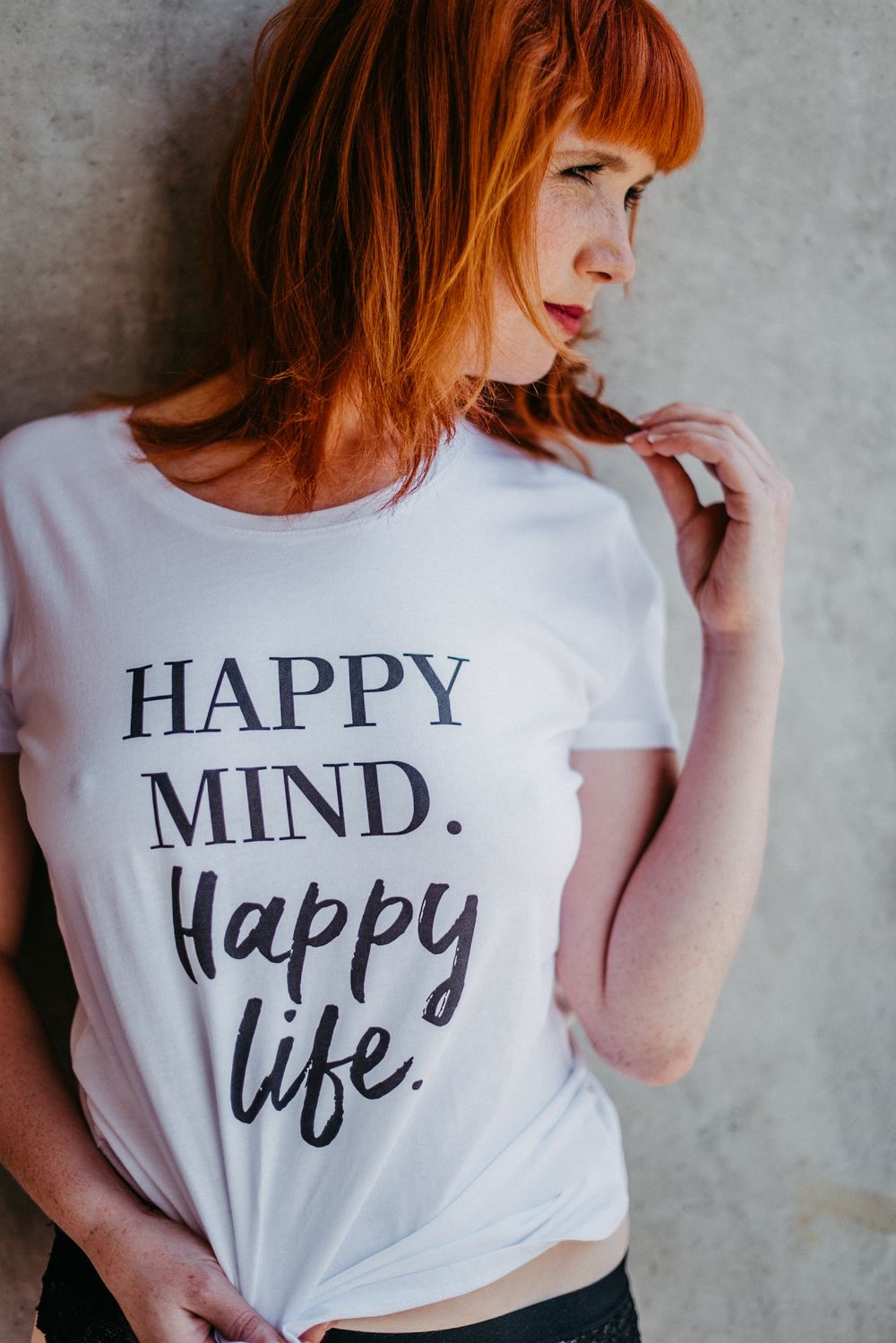 Frau mit rotem Haar trägt ein weißes T-Shirt mit der Aufschrift "Happy Mind. Happy Life." und steht vor einer grauen Wand.