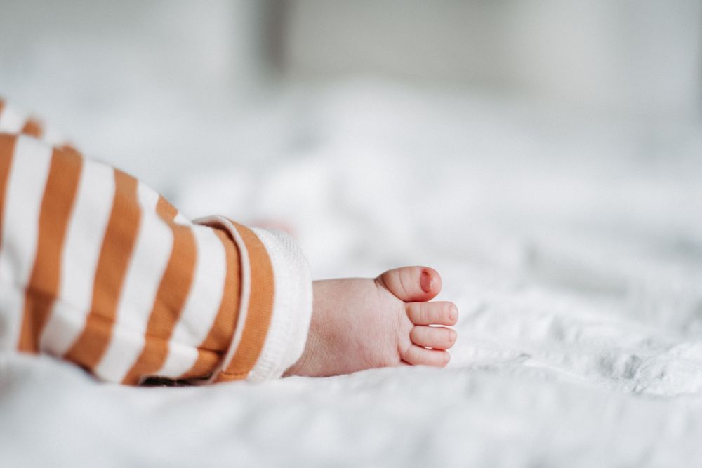 Ein nackter Babyfuß guckt aus einer gestreiften Hose, Das Baby liegt auf einer weissen Decke.