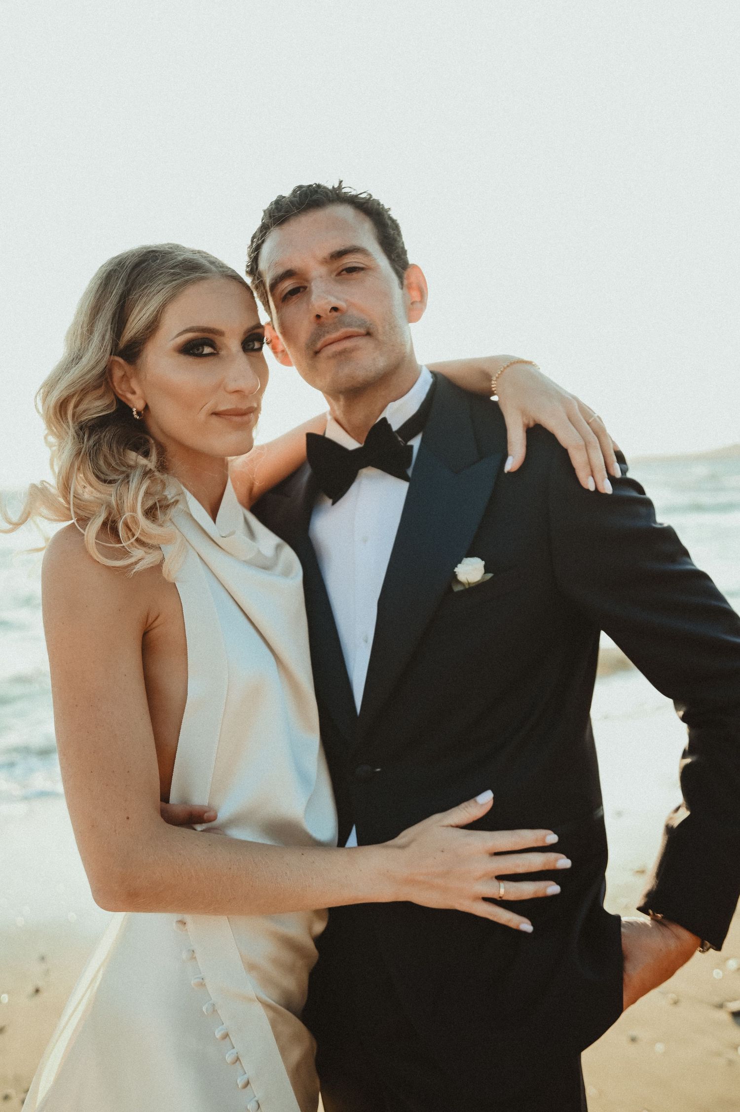 Glamorous couple in formal attire pose for sunset beach wedding portrait.