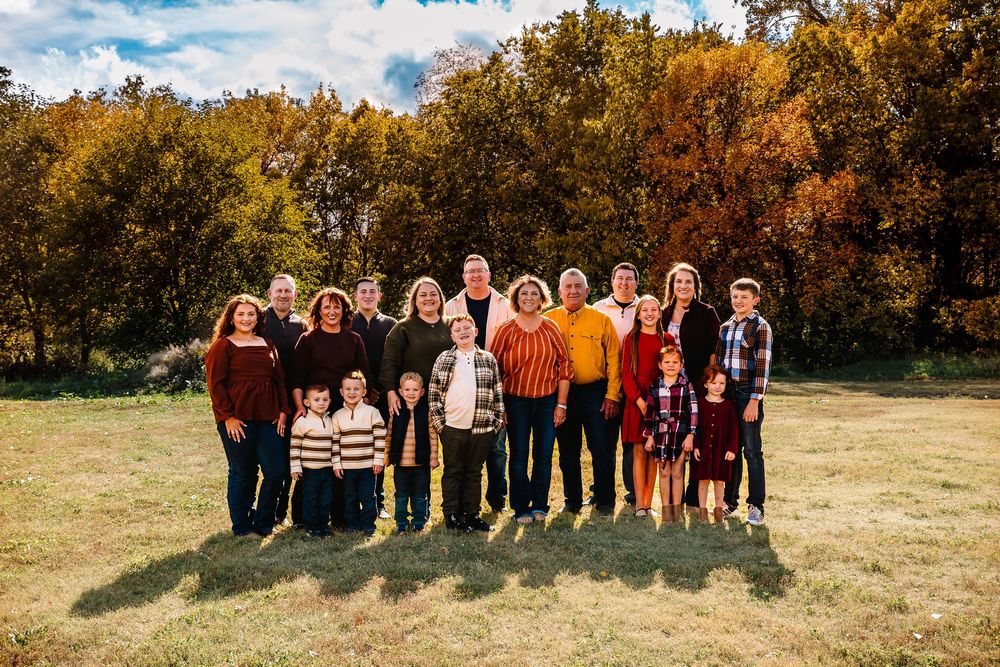 A group of adults and children gathered outdoors in casual wear standing together against a natural fall backdrop.