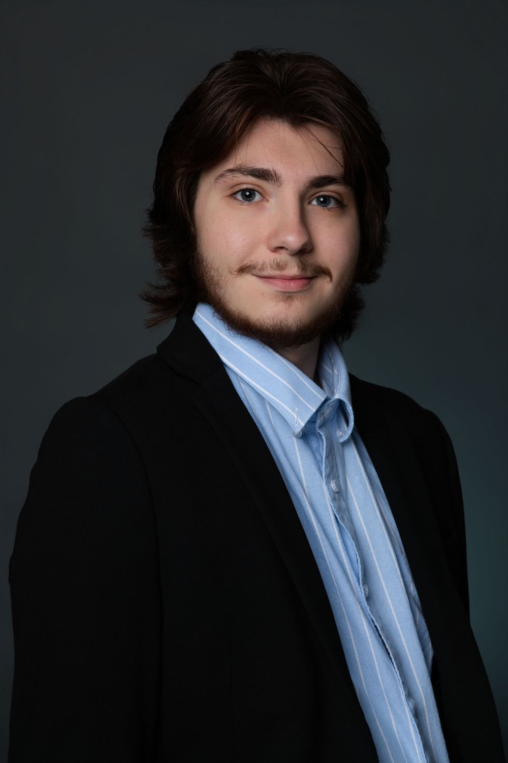 Young man with brown hair in black blazer and blue striped shirt smiling at camera against dark background.