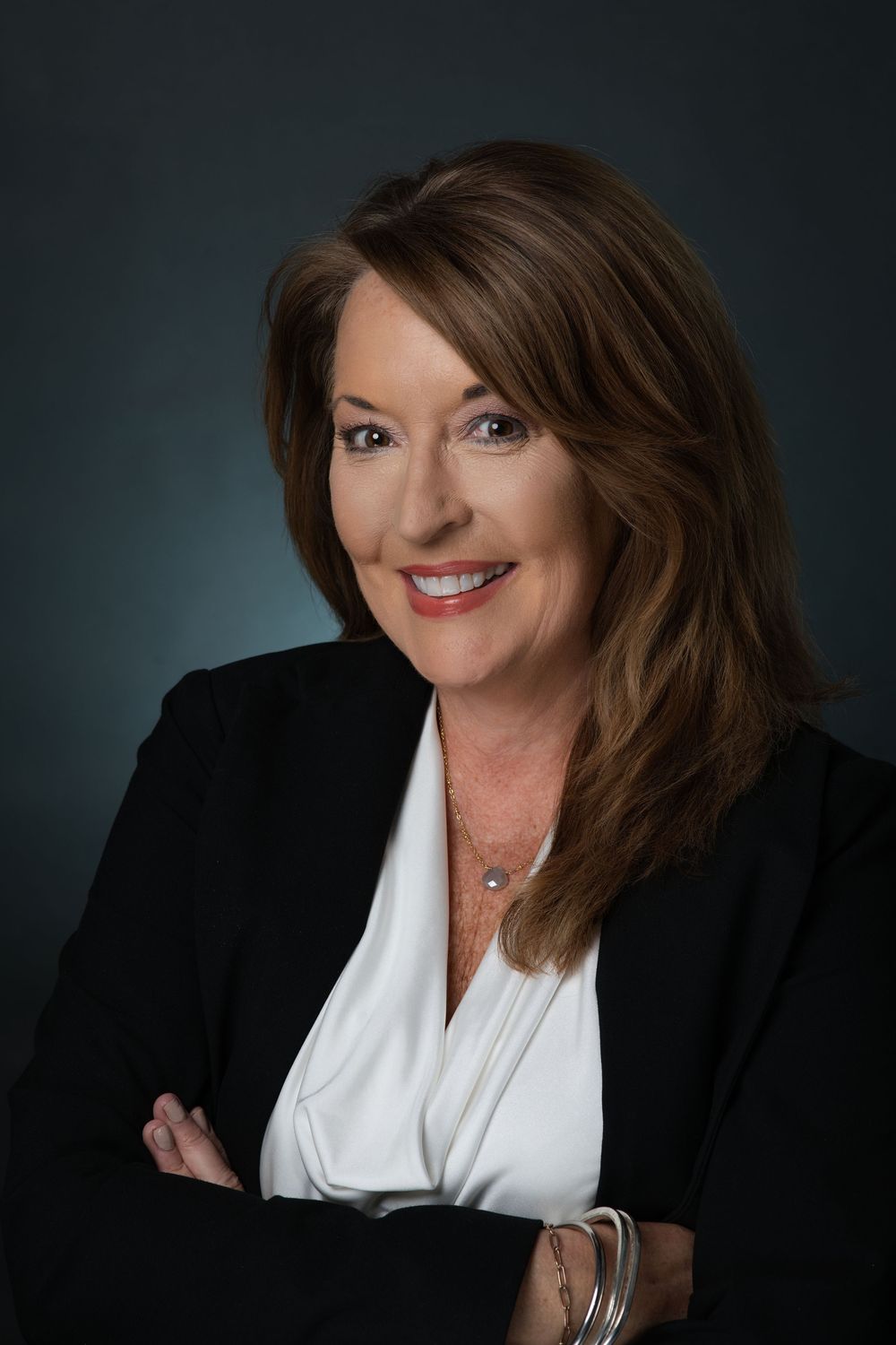 Professional woman in black blazer and white blouse smiling with arms crossed against dark background.
