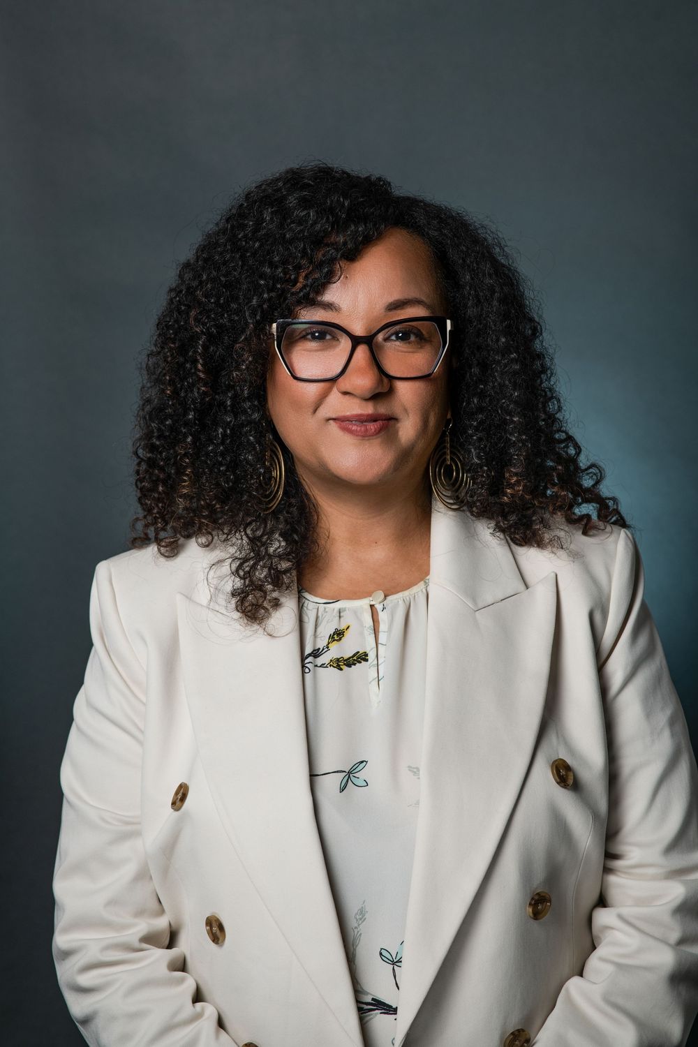Professional woman with curly hair and glasses wearing a white blazer, smiling confidently against a dark background.