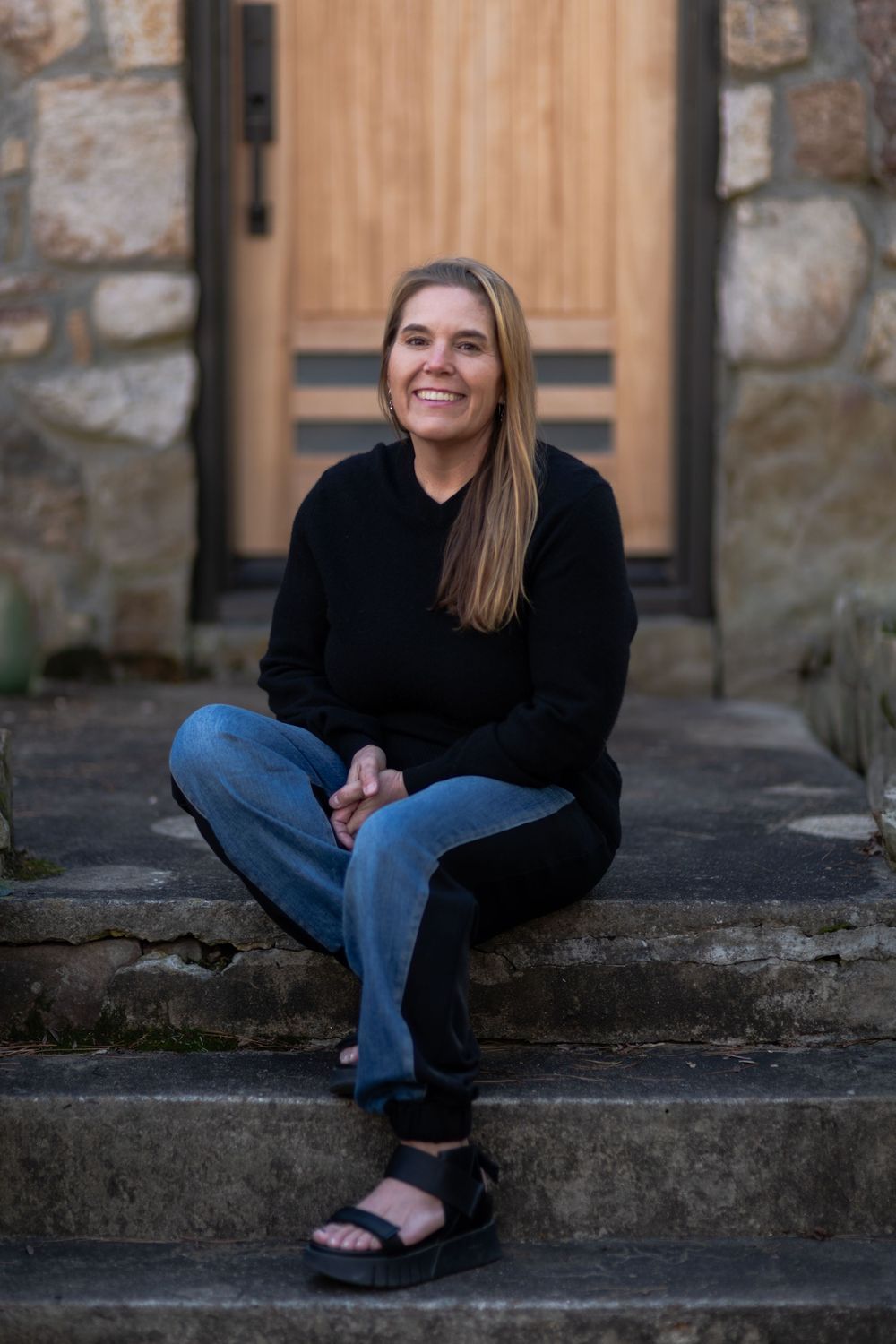Smiling woman in black sweater and jeans sitting on stone steps in front of a wooden door.
