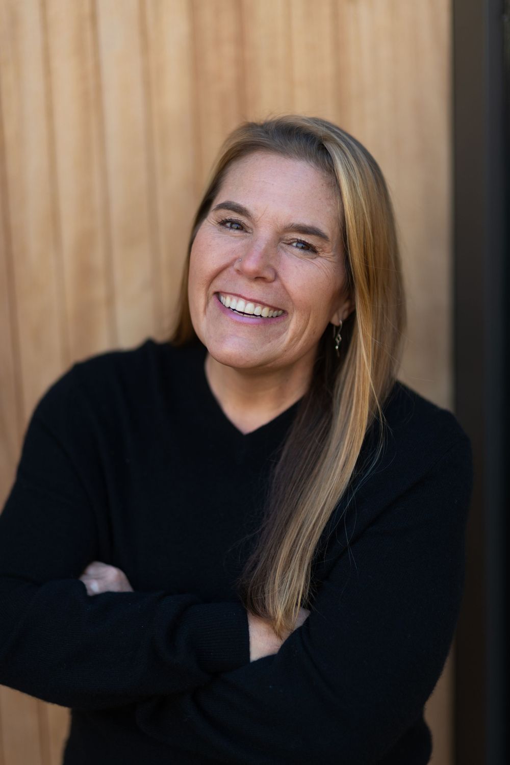 Smiling woman with blonde hair, arms crossed, wearing a black sweater, posing against a wooden background.