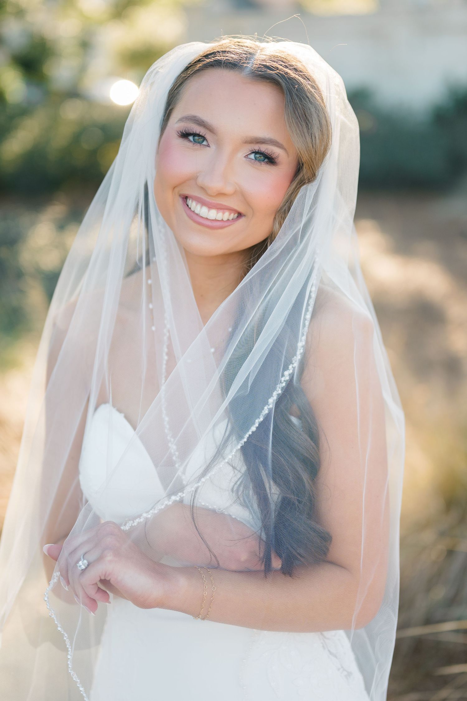 Elegant bride in a white pearl-embellished wedding gown with sheer sleeves holds a pastel bouquet in outdoor setting.