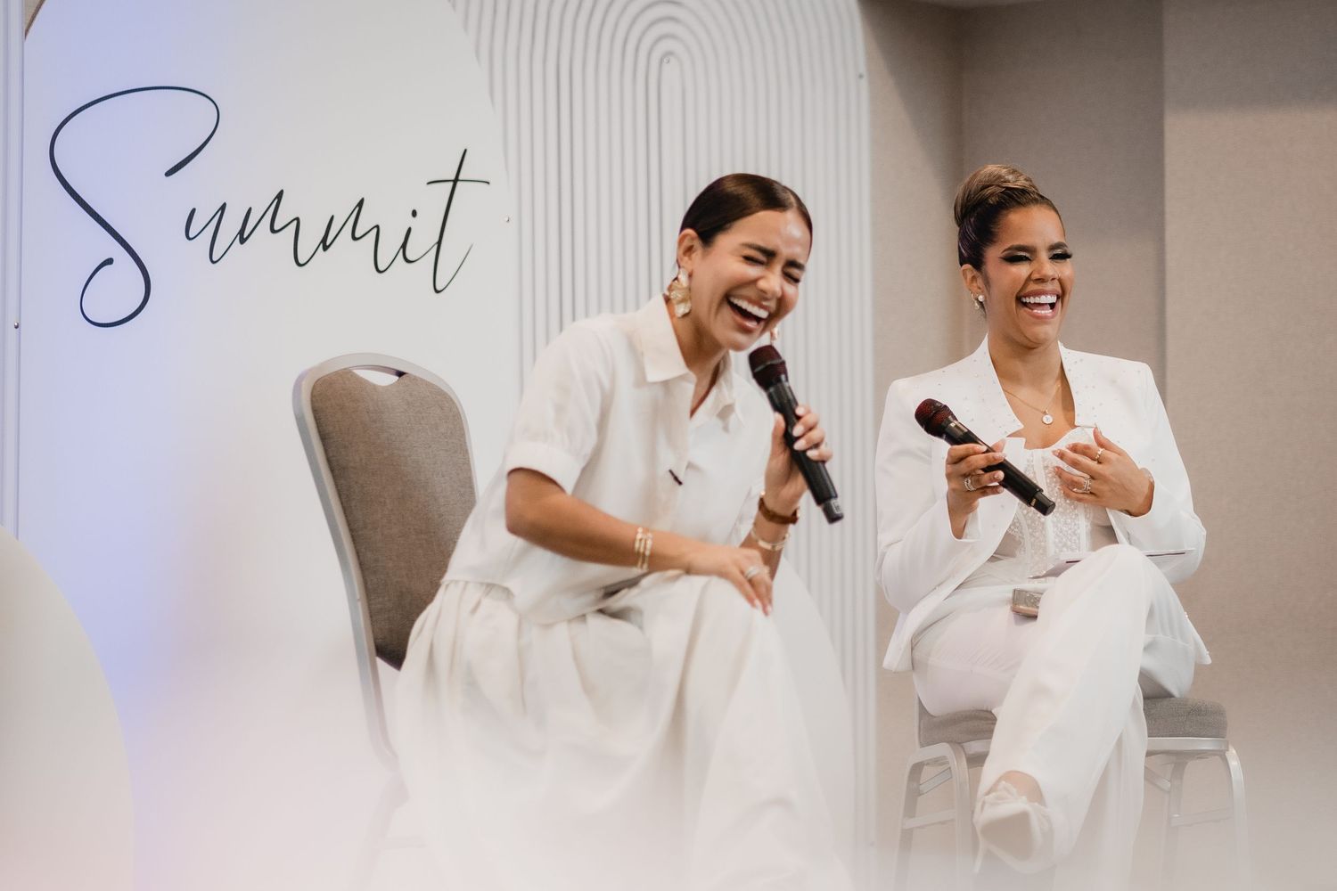 Two people dressed in white outfits sit on chairs laughing and speaking into microphones during a summit event.