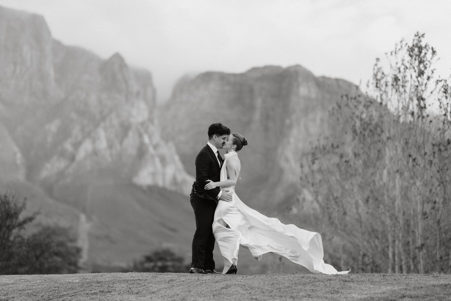 A couple in wedding attire silhouetted against dramatic mountain peaks and a cloudy sky in a scenic outdoor landscape.