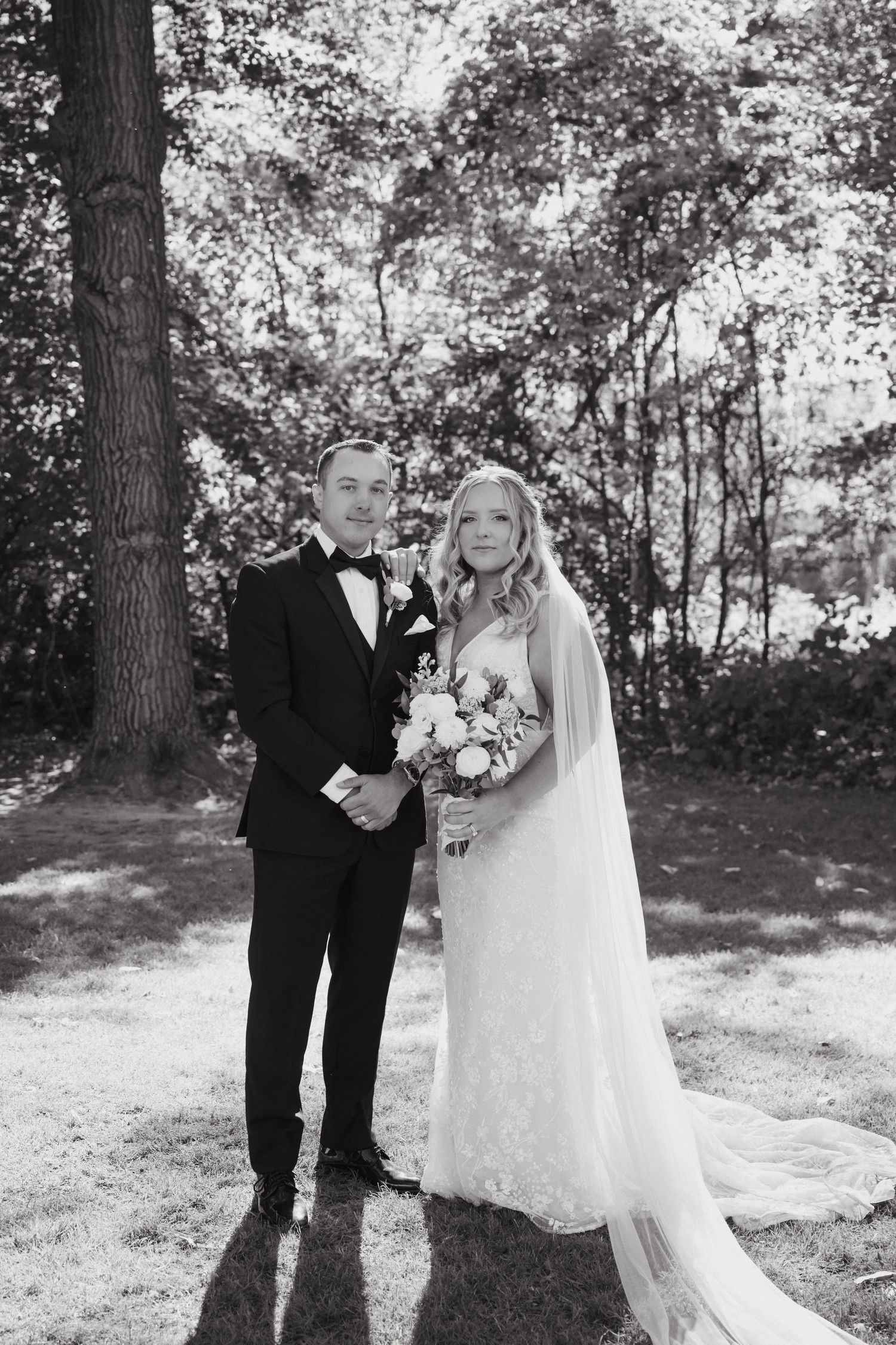 Classic black and white wedding portrait of newlyweds standing together in wooded setting.