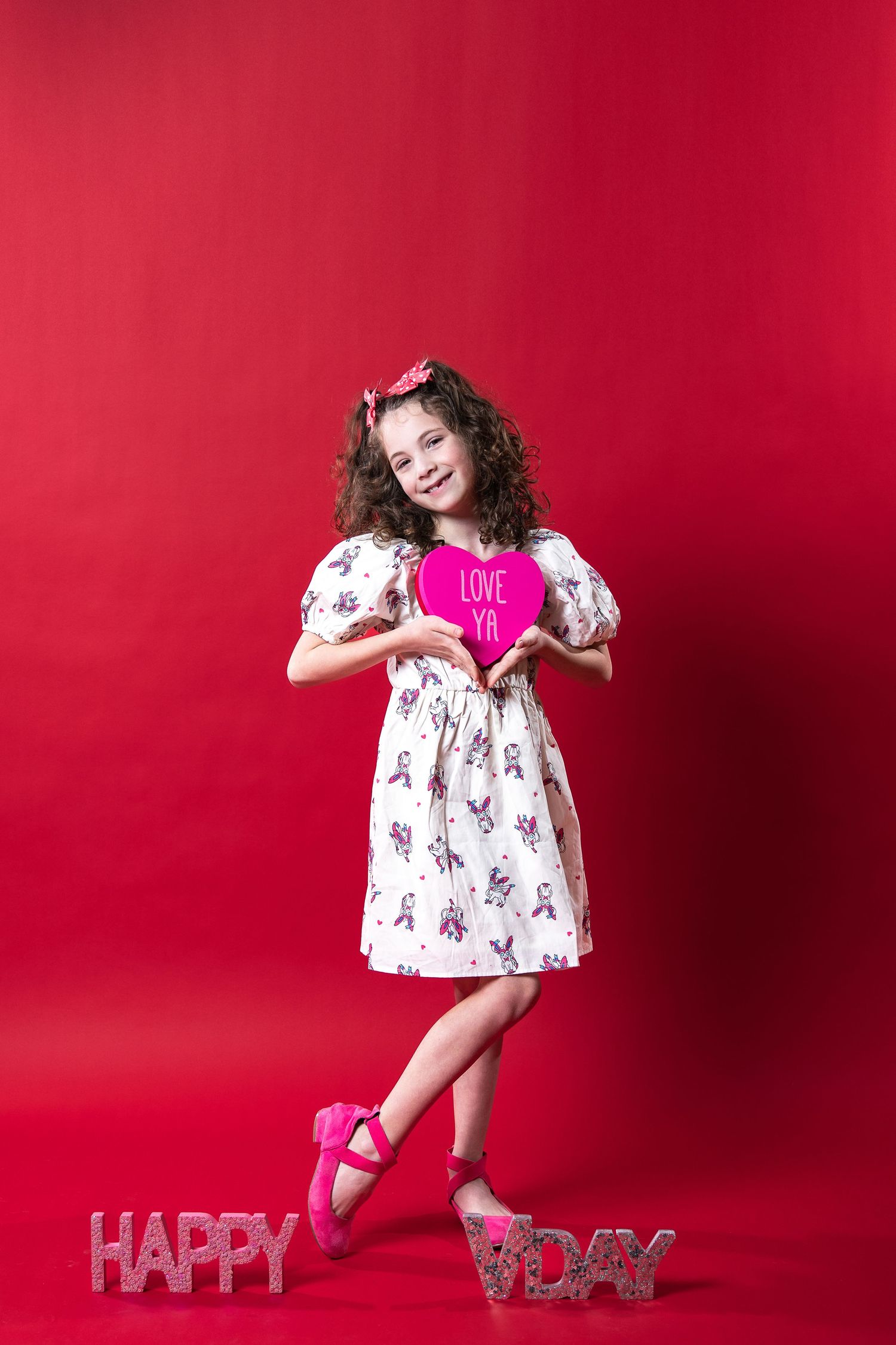 Child in floral dress holding pink heart against red background with "Happy Day" text decoration.