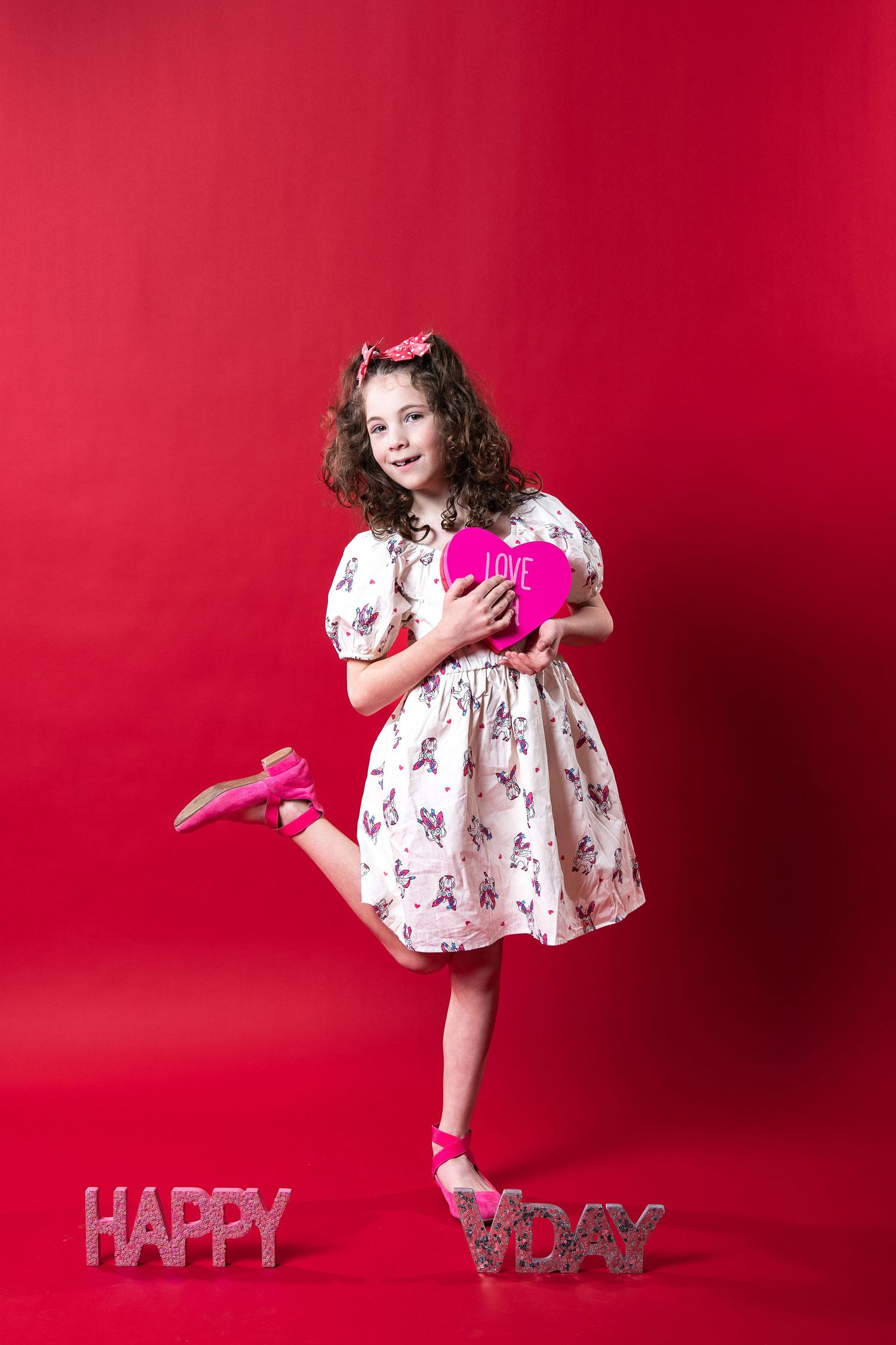Young person in floral dress holding pink heart stands on one foot against red backdrop with 'HAPPY' text decoration.