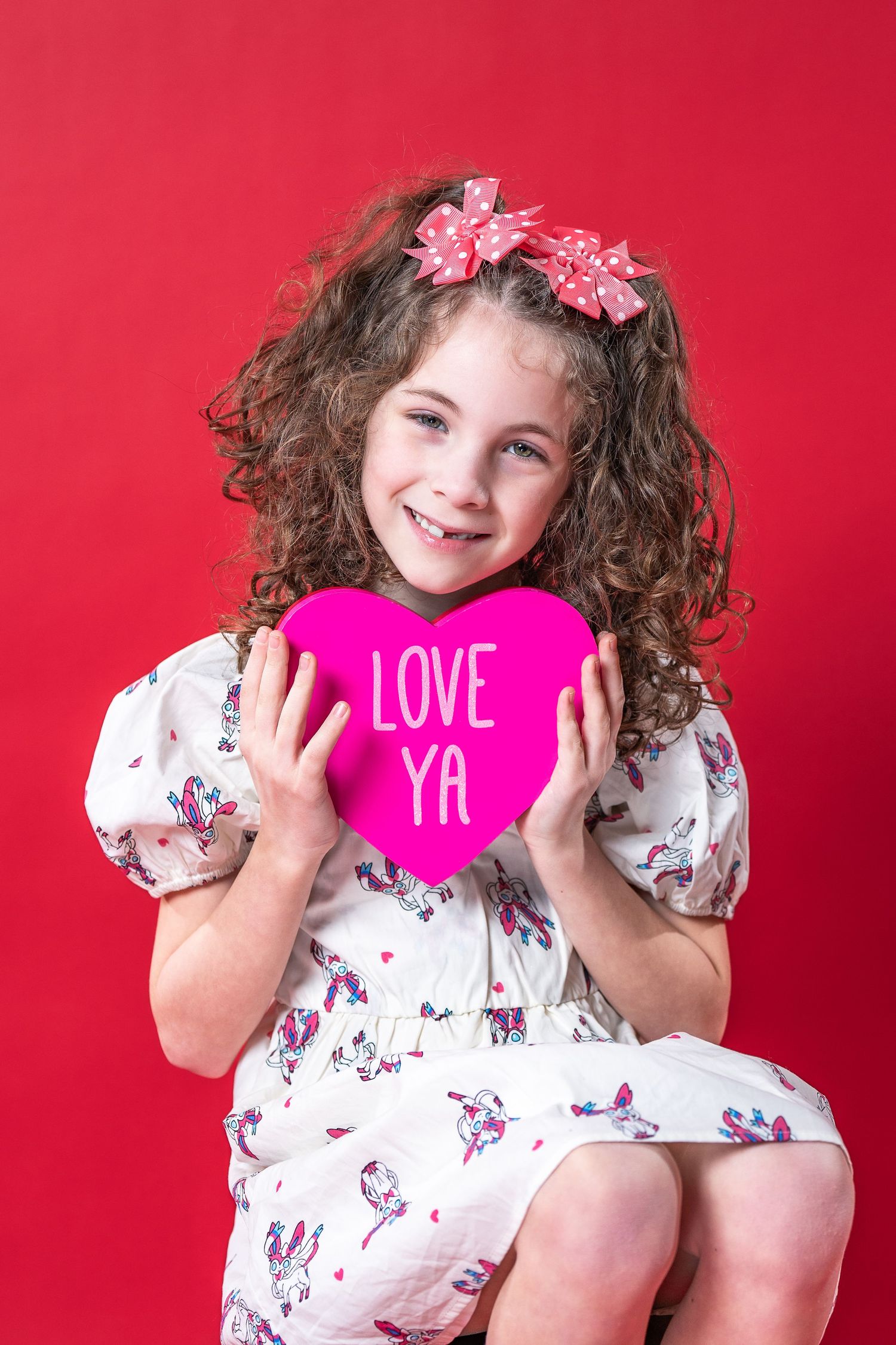 Young child with curly hair in floral dress holding pink heart shape with 'LOVE YA' text against red background.
