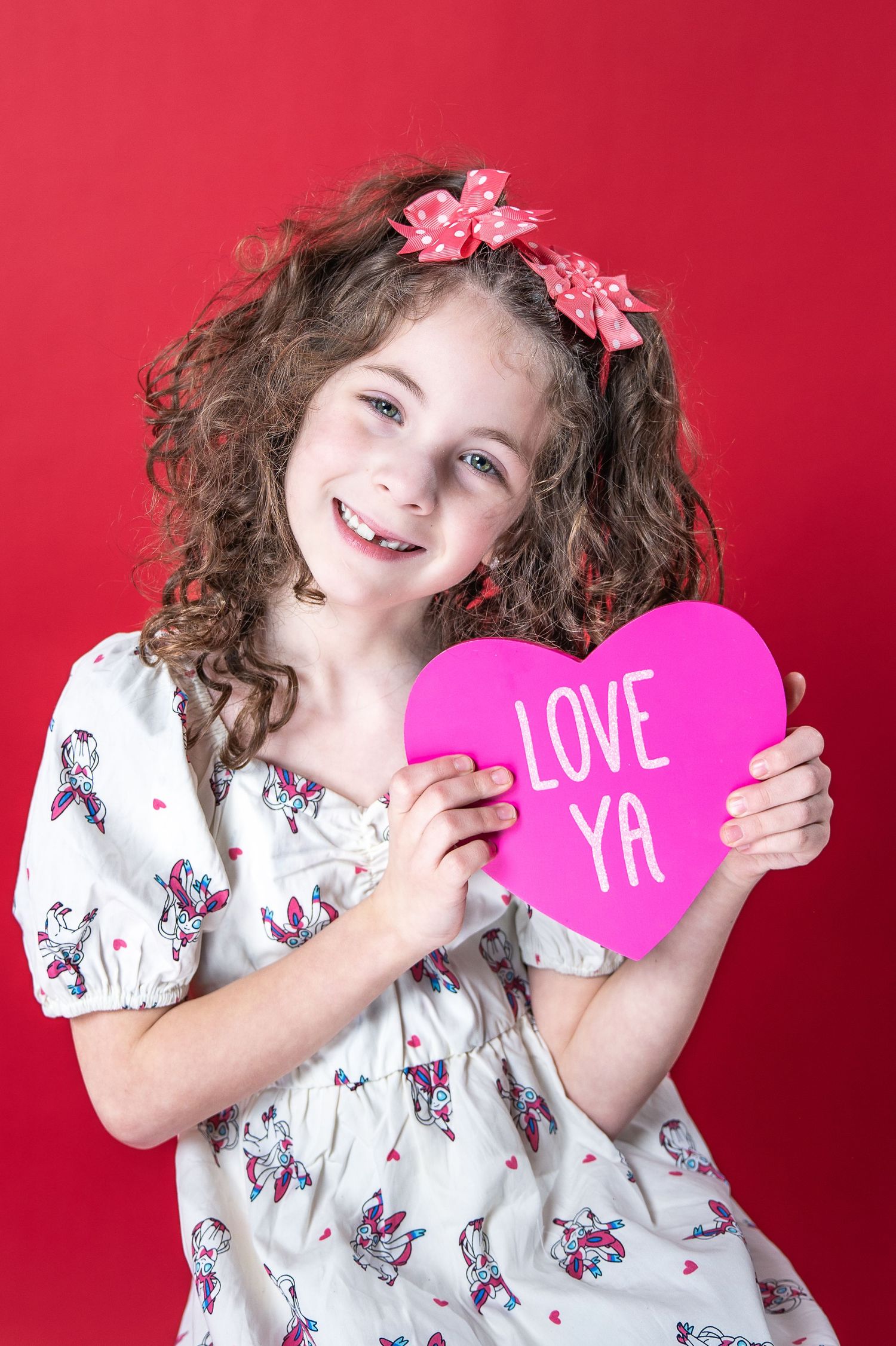 Child with curly hair and pink bows holding a pink heart that says 'LOVE YA' against a red background.