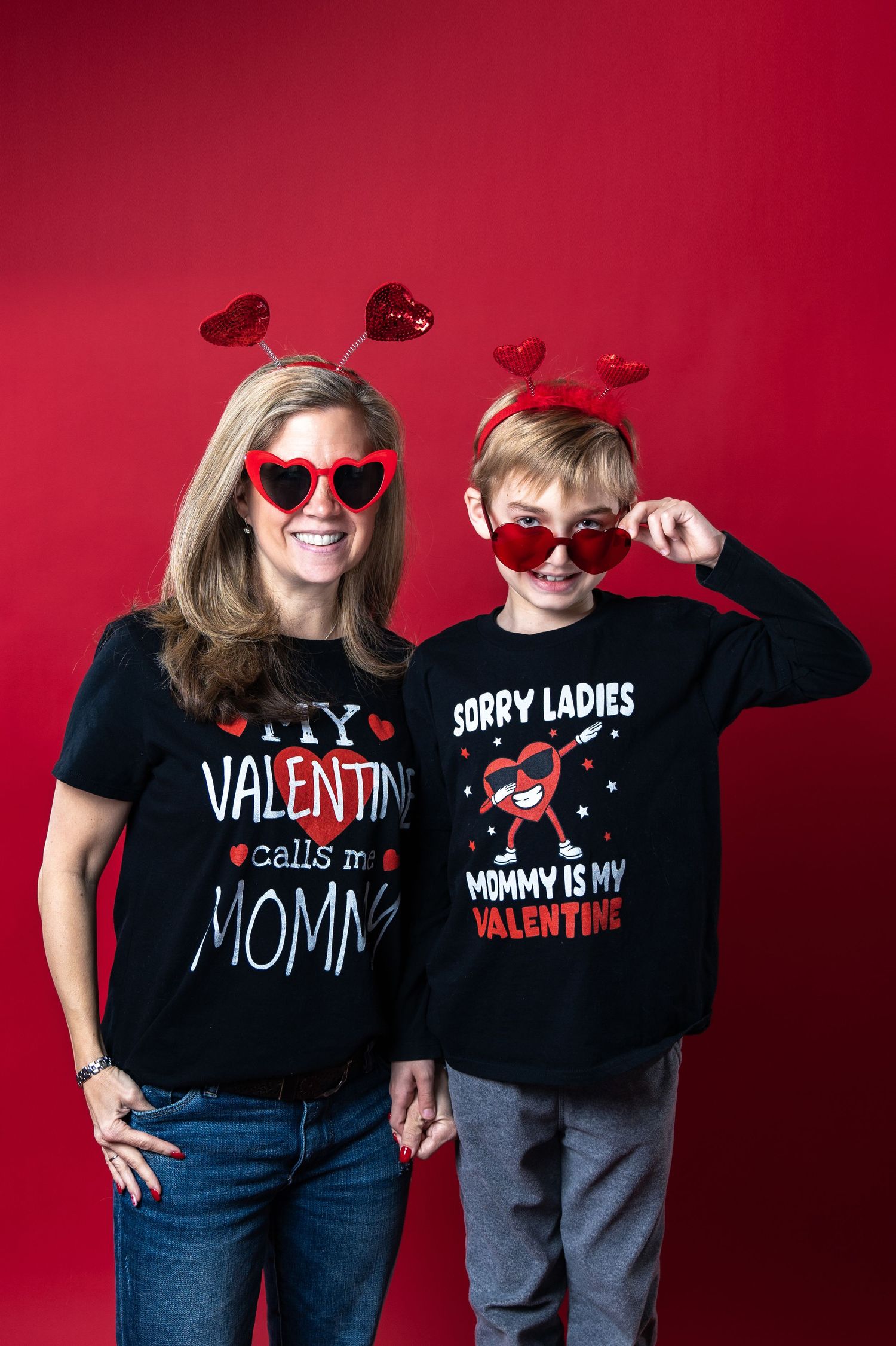 A parent and child wearing matching Valentine's Day themed black t-shirts and heart headbands against a red background.