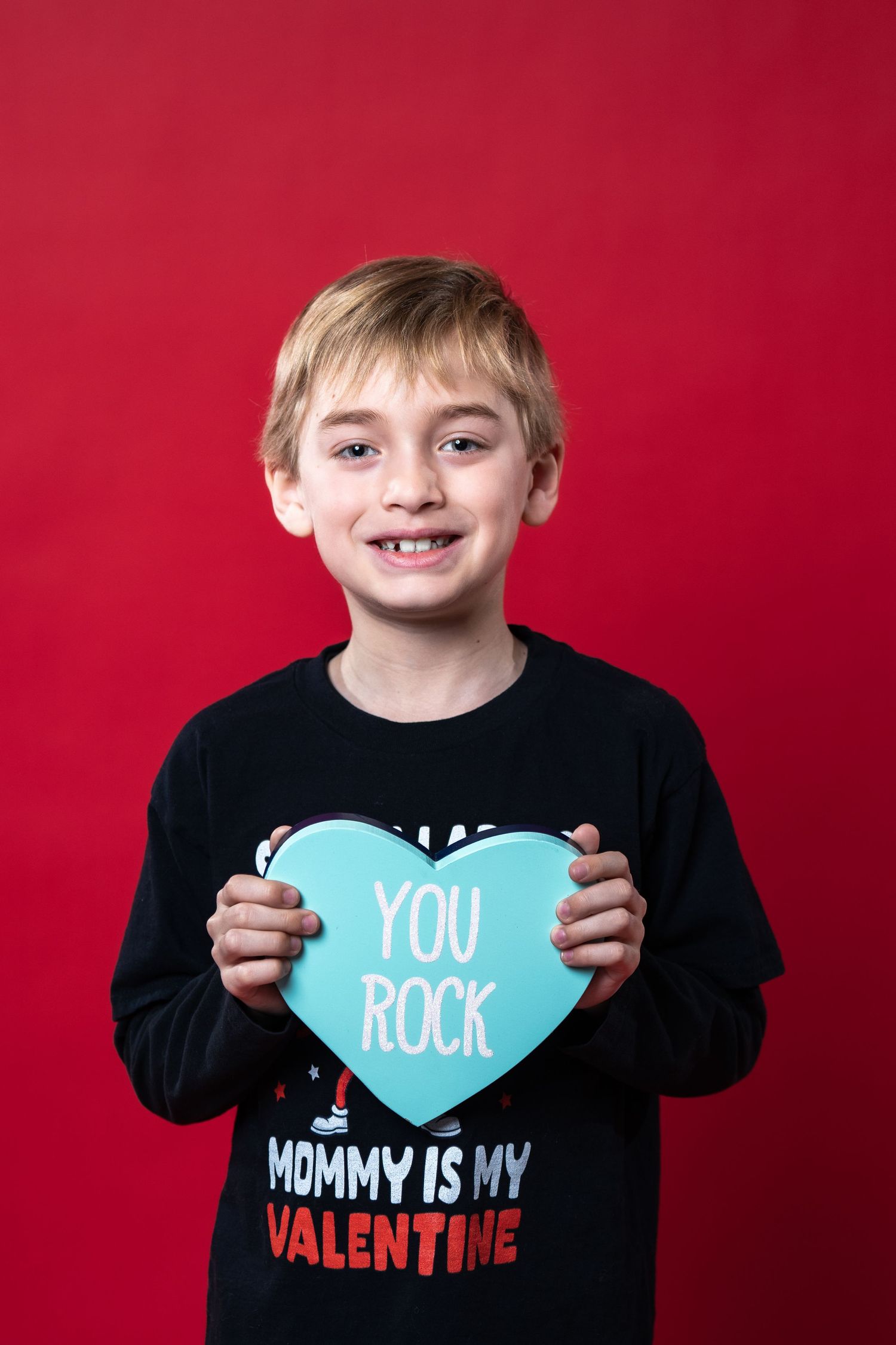 Child wearing "Mommy is my Valentine" shirt holds light blue heart shape against red background while smiling.