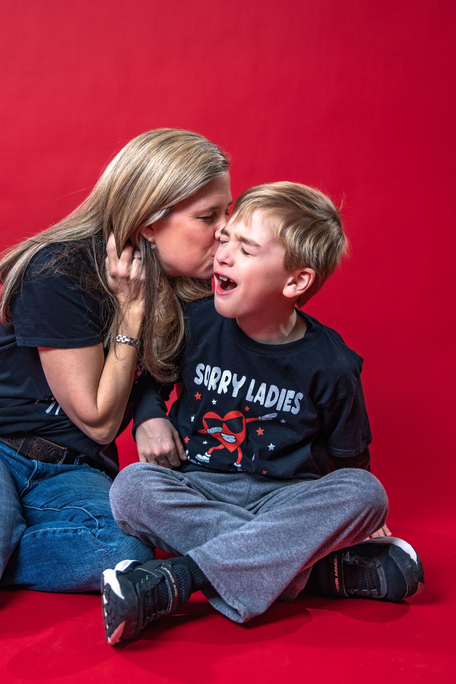A young boy wearing a "Sorry Ladies" t-shirt reacts playfully while someone whispers in his ear against a red backdrop.