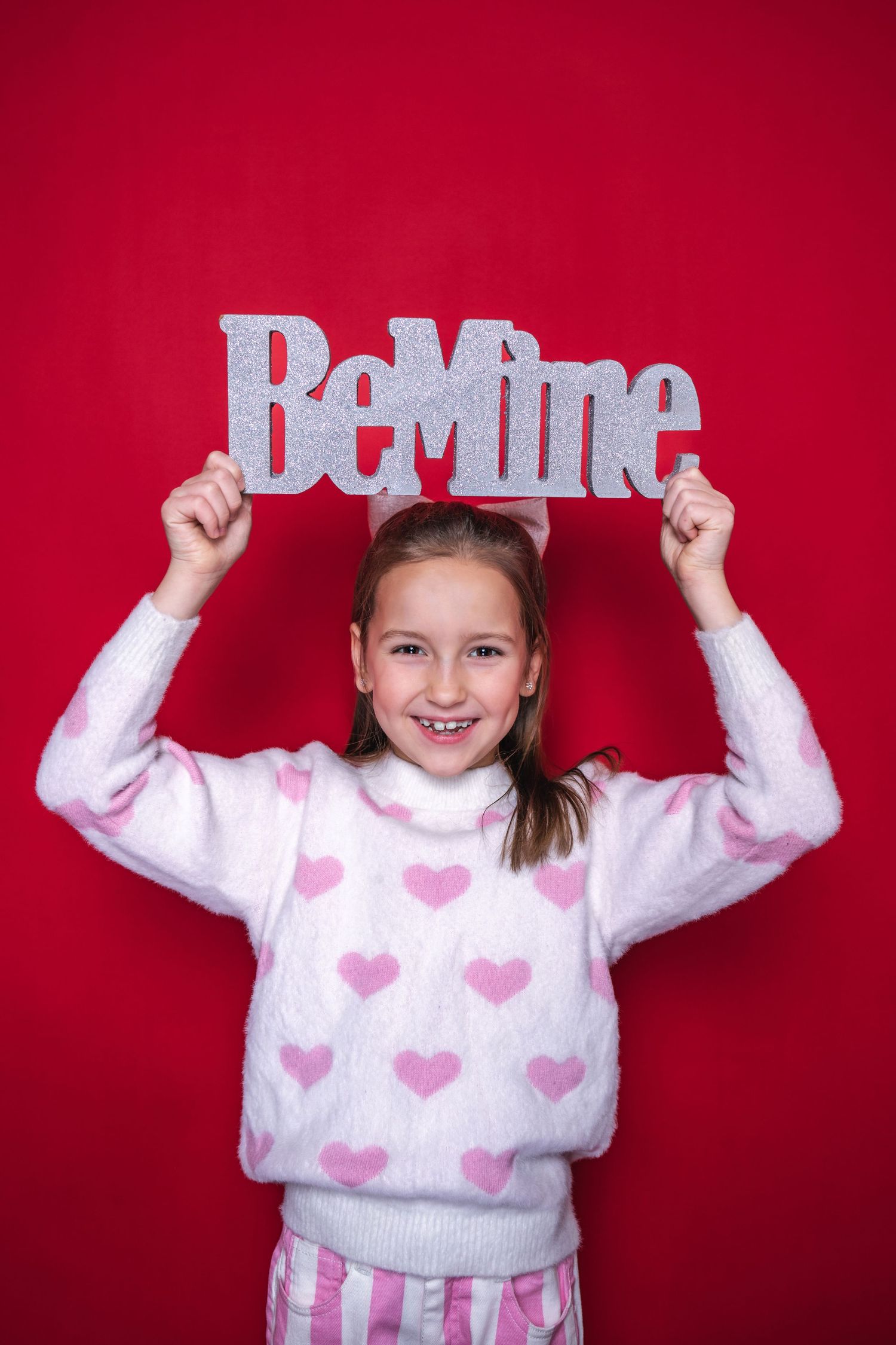 A child wearing a pink heart sweater holds up a 'Be Mine' text cutout against a red background.
