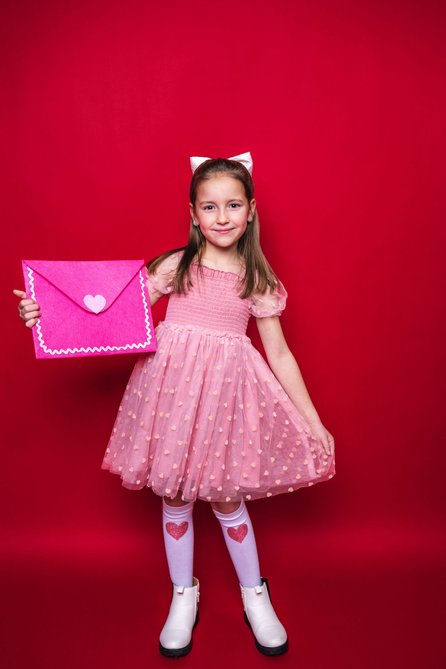 A child in a pink polka dot dress and white stockings holds up a large pink envelope against a red background.
