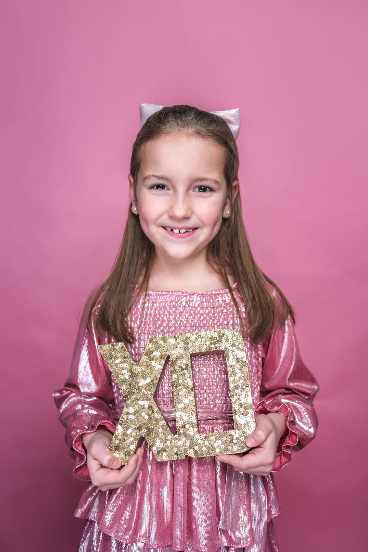 A young child in a pink velvet dress poses with a gold glittery XO decoration against a pink backdrop.
