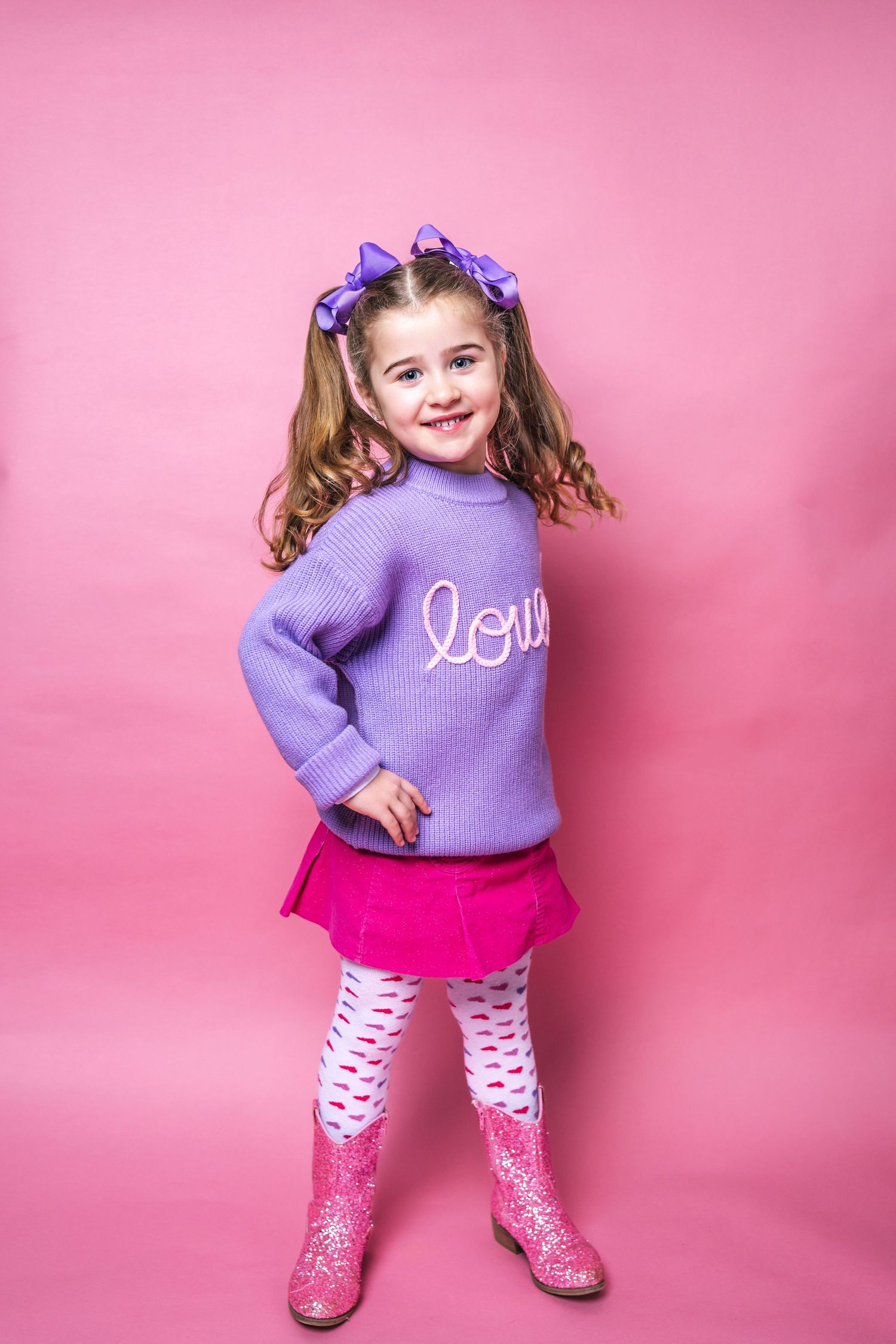 A young child wearing a purple 'love' sweater, pink skirt, heart leggings and glittery boots poses against a pink background.