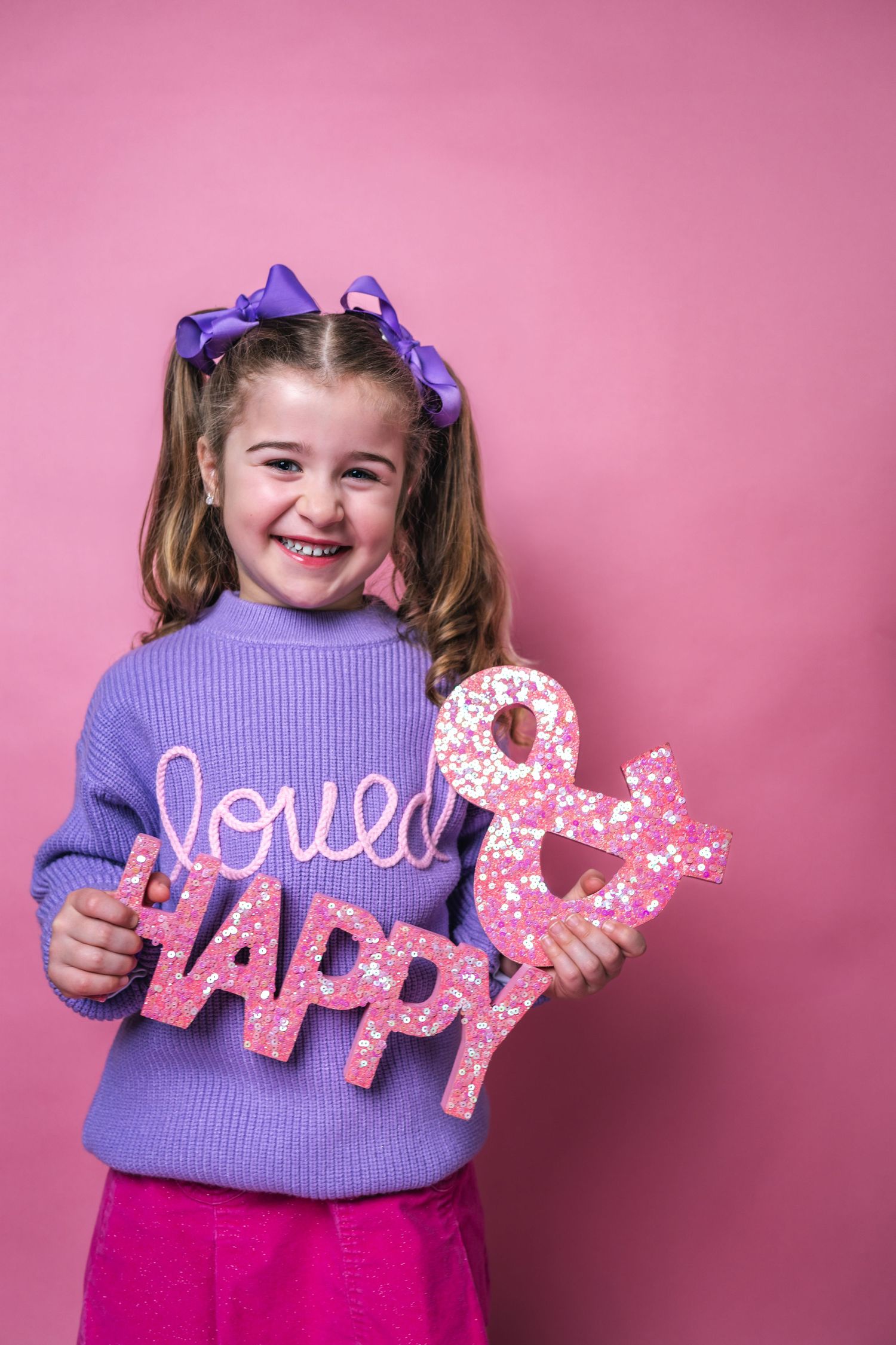 Child in purple sweater holds decorative 'HAPPY' and 'LOVE' letters against pink background.
