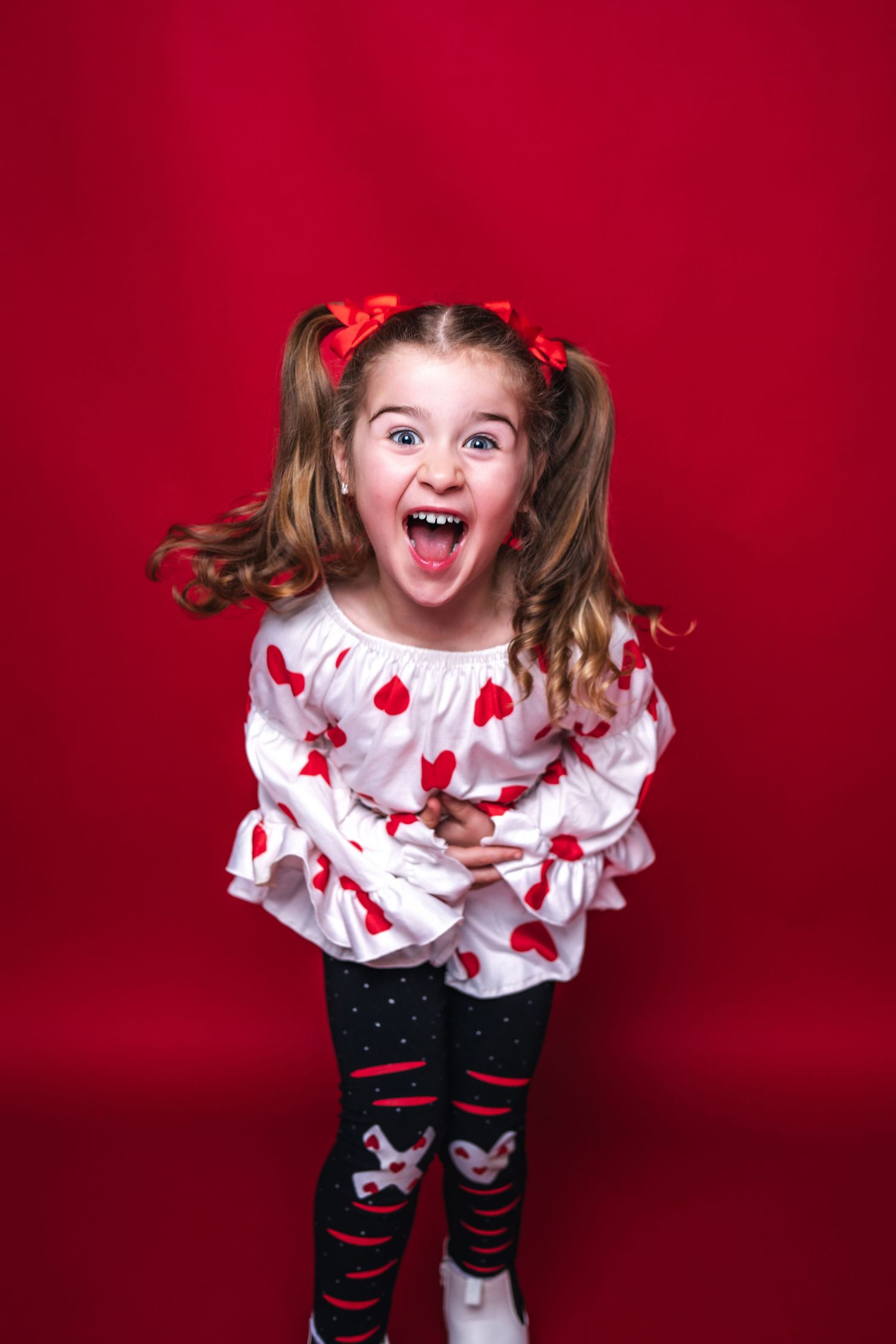 An excited child wearing a heart-patterned sweater and patterned leggings poses energetically against a red background.